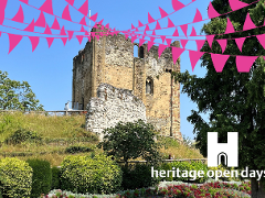 A medieval castle ruin rises above landscaped gardens, decorated with bright pink bunting against a clear blue sky. Sunlit stone walls and a historic tower stand on a grassy hill, while colourful flowerbeds and mature trees frame the scene. The Heritage O